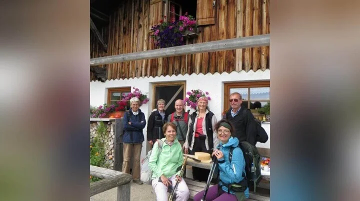 Wandergruppe vor der Rinner Alm | © DAV Markt Schwaben | Foto Erwin Matzinger Acht Wandernde stehen vor der Rinner Alm mit Holzfassade und Blumen; eine Person sitzt mit Käseteller auf der Bank. | © DAV Markt Schwaben | Foto Erwin Matzinger
