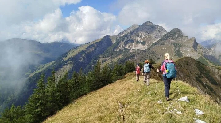 Ankunft an der Wildenkaralm | © DAV Markt Schwaben | Foto Helga Peters Drei Wandernde stehen vor rustikaler Almhütte aus Holz und Stein; hügelige Landschaft und Wolken im Hintergrund. | © DAV Markt Schwaben | Foto Helga Peters