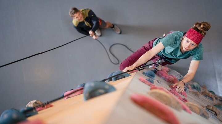 Frau klettert an einer Indoor-Kletterwand, gesichert von einem Mann im Hintergrund. Beide sind in Kletterausrüstung. | © DAV/Marisa Koch 2023