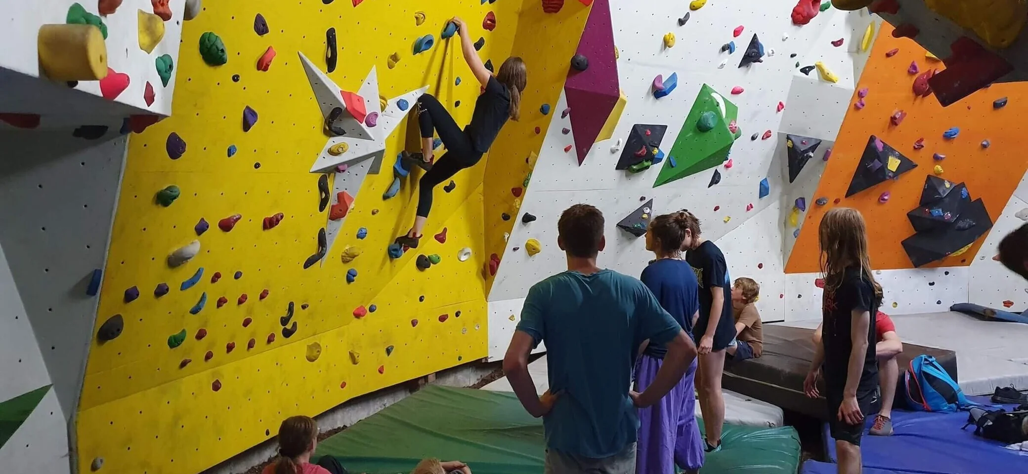 Jugendgruppe Kletter- und Bouldergruppe | © DAV Markt Schwaben Jugendliche in einer Kletterhalle erleben Teamgeist und Abenteuer: Ein junger Kletterer erklimmt eine Wand mit farbigen Griffen, während andere auf Matten sitzen und zuschauen. Die Szene spiegelt den Spaß und die Gemeinschaft der Jugendgruppe wider. | © DAV Markt Schwaben